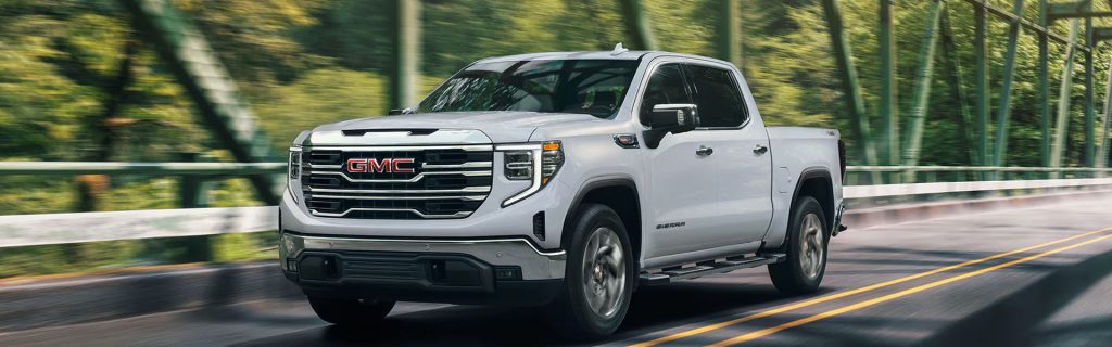 A white GMC Sierra truck driving across a bridge surrounded by greenery.